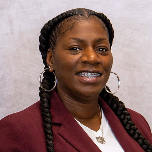 Woman with long braids, wearing a maroon blazer, white shirt, silver hoop earrings, and braces, smiling in front of a light gray background.