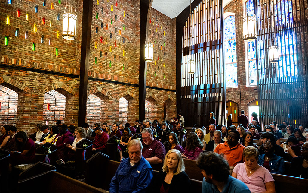 People sit in pews inside a brick-walled church with stained glass windows, colorful wall accents, and hanging light fixtures during a gathering.