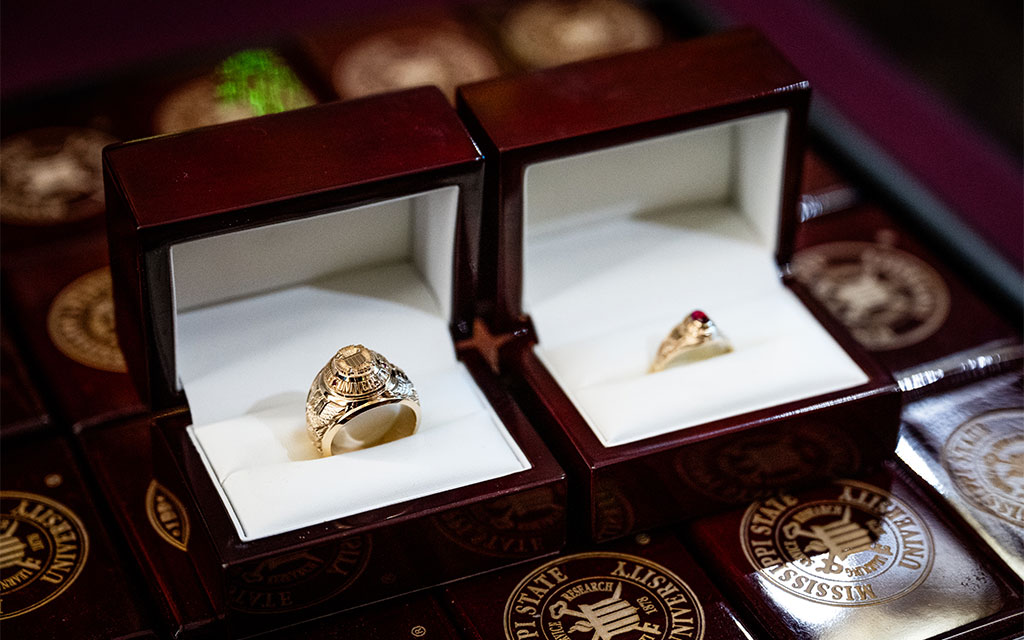 Two rings in wooden boxes: one is a large gold class ring, the other a smaller gold ring with a red stone. Both boxes display a university emblem inside the lid.