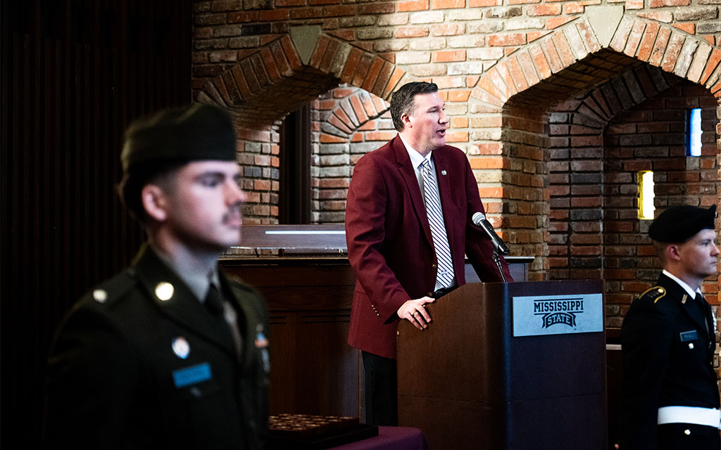 A man speaks at a podium labeled “Mississippi State,” while two uniformed military personnel stand in the foreground. Brick walls with arched openings are visible behind the speaker.