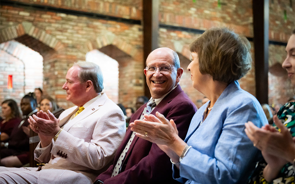 Three adults in formal attire sit and clap in an indoor event space with exposed brick walls, while others watch in the background.