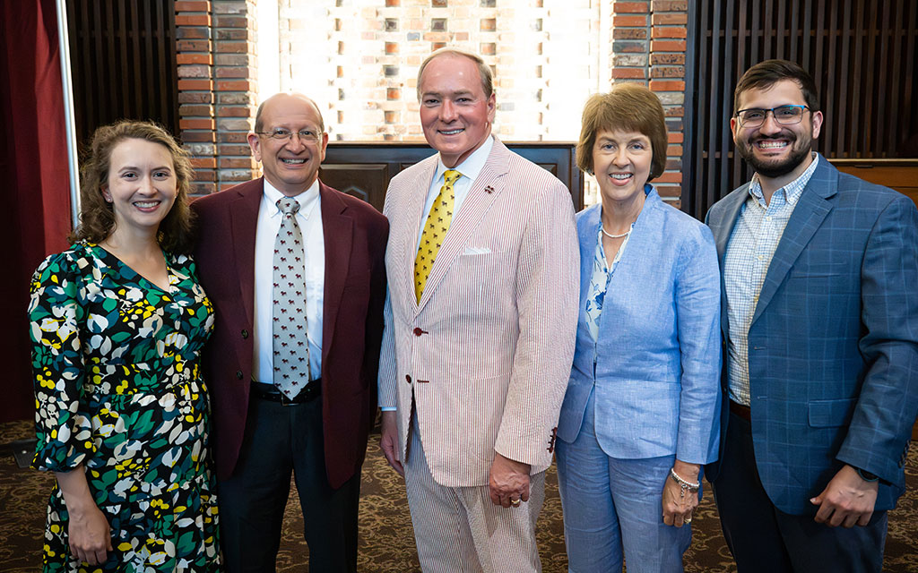 Five adults in formal attire stand side by side indoors, smiling at the camera with a brick wall and window behind them.