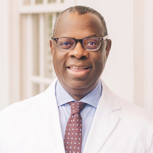 Man wearing glasses, a white lab coat, a light blue shirt, and a patterned tie, standing indoors in front of a light-colored background.