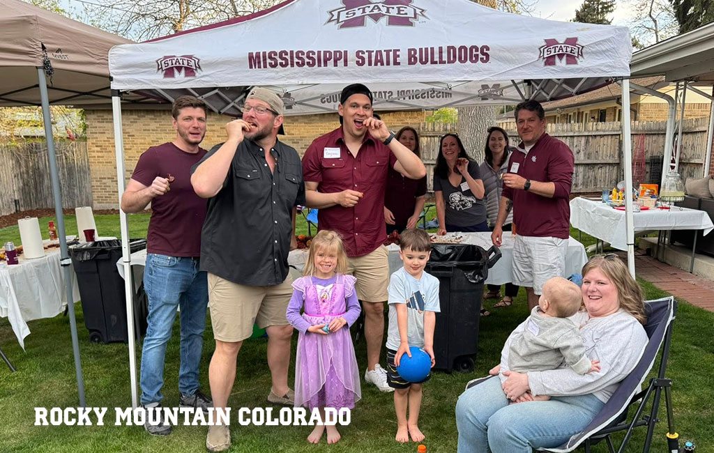 A group of adults and children pose and socialize under a Mississippi State Bulldogs tent at an outdoor gathering in a backyard.