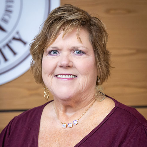 A woman with short, light brown hair and blue eyes is smiling at the camera. She is wearing a maroon top, a necklace, and dangling earrings, with a partial university seal in the background.