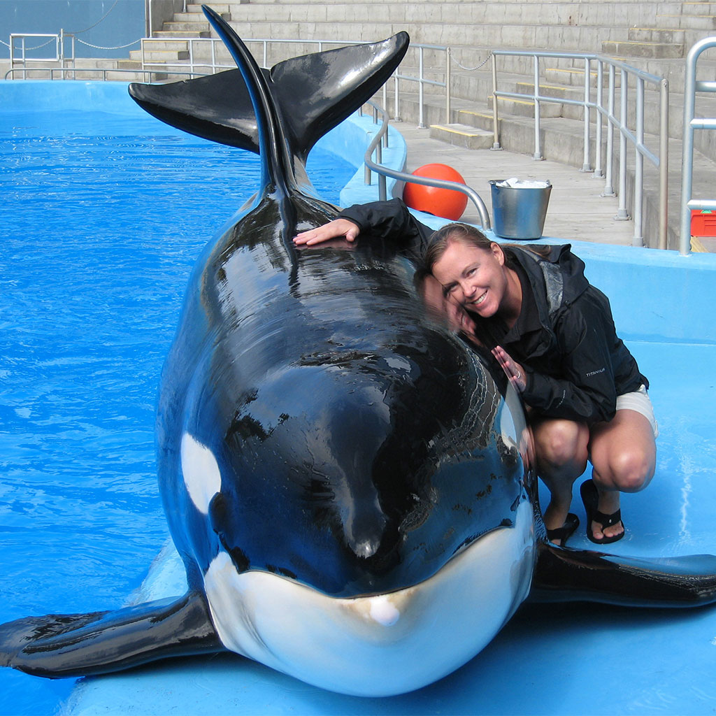 A woman crouches beside a large orca in a pool, smiling and touching the whale’s back. Steps, rails, and pool equipment are visible in the background.