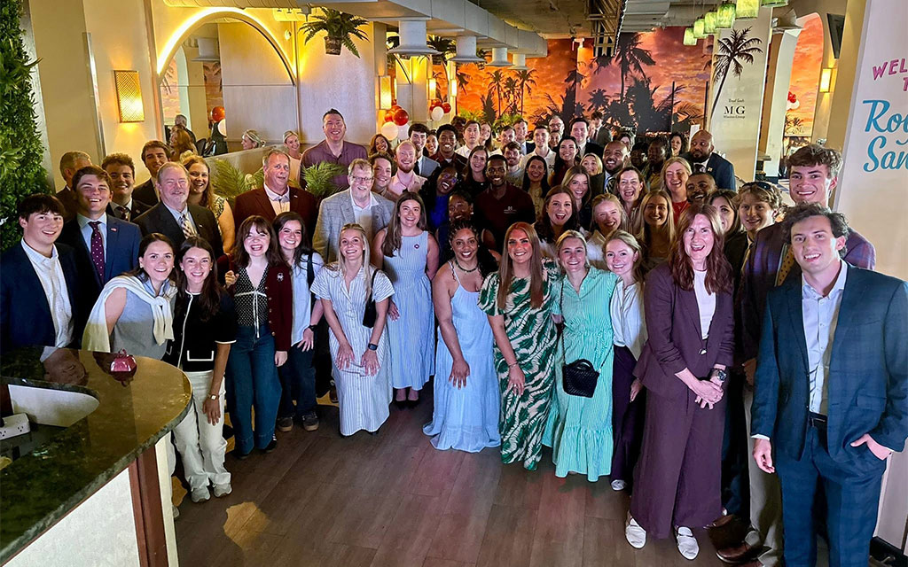 A large group of people, dressed in semi-formal to formal attire, gather indoors for a group photo at an event with festive decorations and a tropical mural in the background.