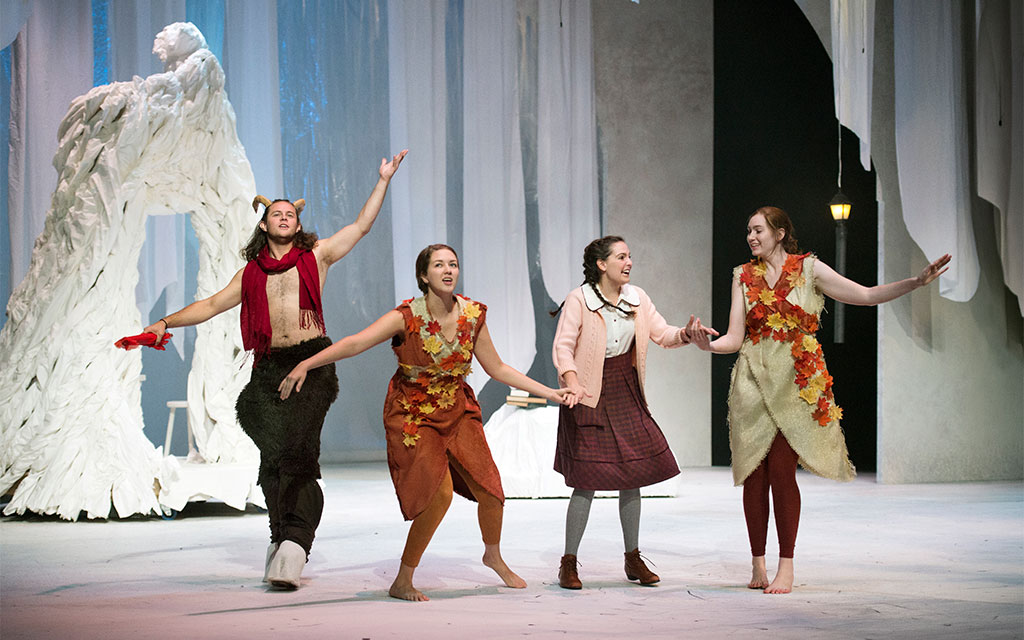 Four actors in costume perform on a snowy stage with white drapery; two wear faun outfits with leaves, and two wear skirts and cardigans, holding hands and singing.