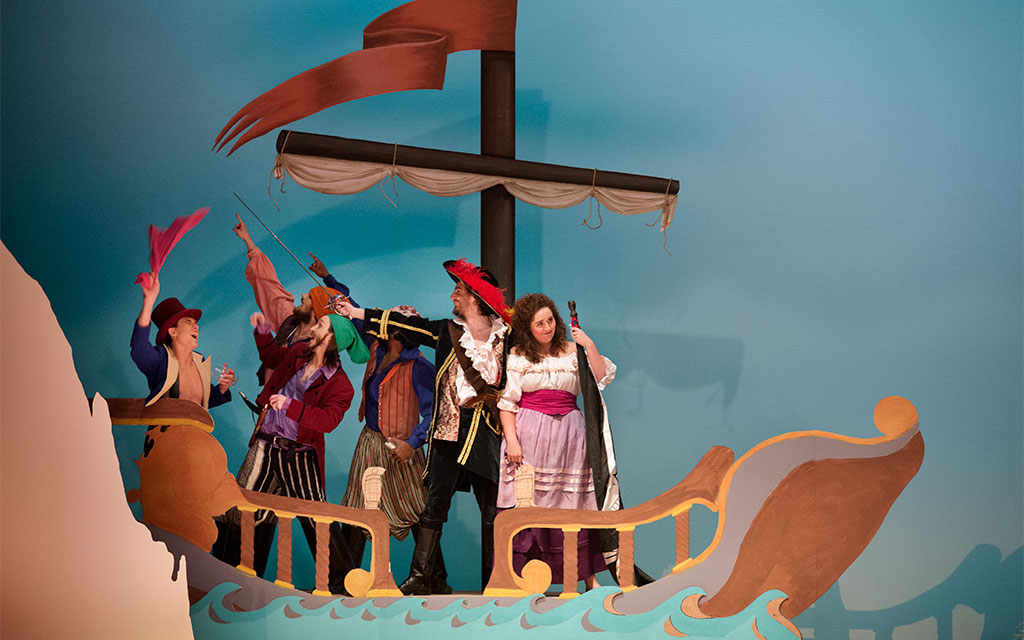Five people dressed as pirates stand on a stage set designed to look like a wooden ship, holding props and wearing colorful costumes, with a blue backdrop.