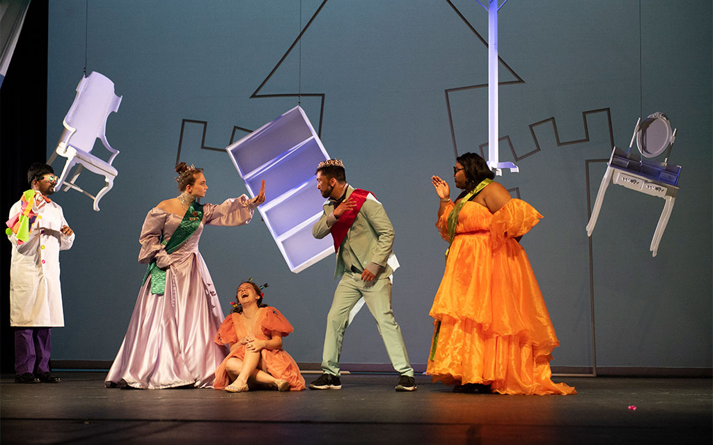 Five actors in colorful costumes perform dramatically on stage with upside-down chairs and a castle backdrop.