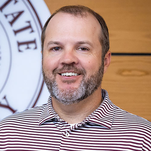 A man with a short beard and striped polo shirt smiles, standing indoors in front of a partial emblem and wood-paneled wall.