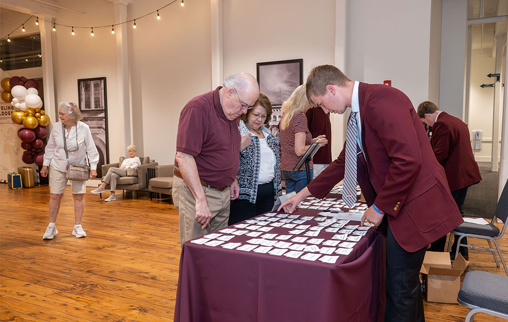 People look through nametags on a table at an indoor event; a man in a maroon jacket assists. Balloons and chairs are visible in the background.