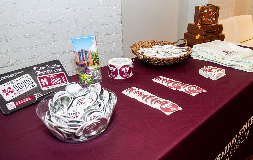A maroon tablecloth displays magnets, stickers, buttons, brochures, napkins, and pens, all featuring Mississippi State University branding, with a white brick wall in the background.