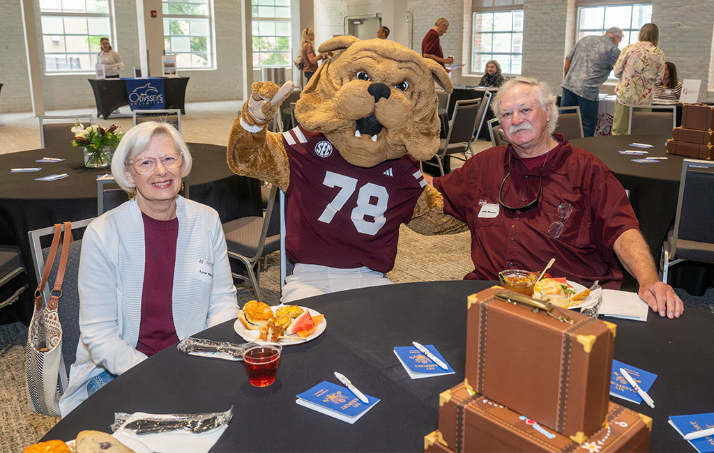 Two people sit at a round table with food and drink, posing with a bulldog mascot in a maroon jersey numbered 78 at an indoor social event.