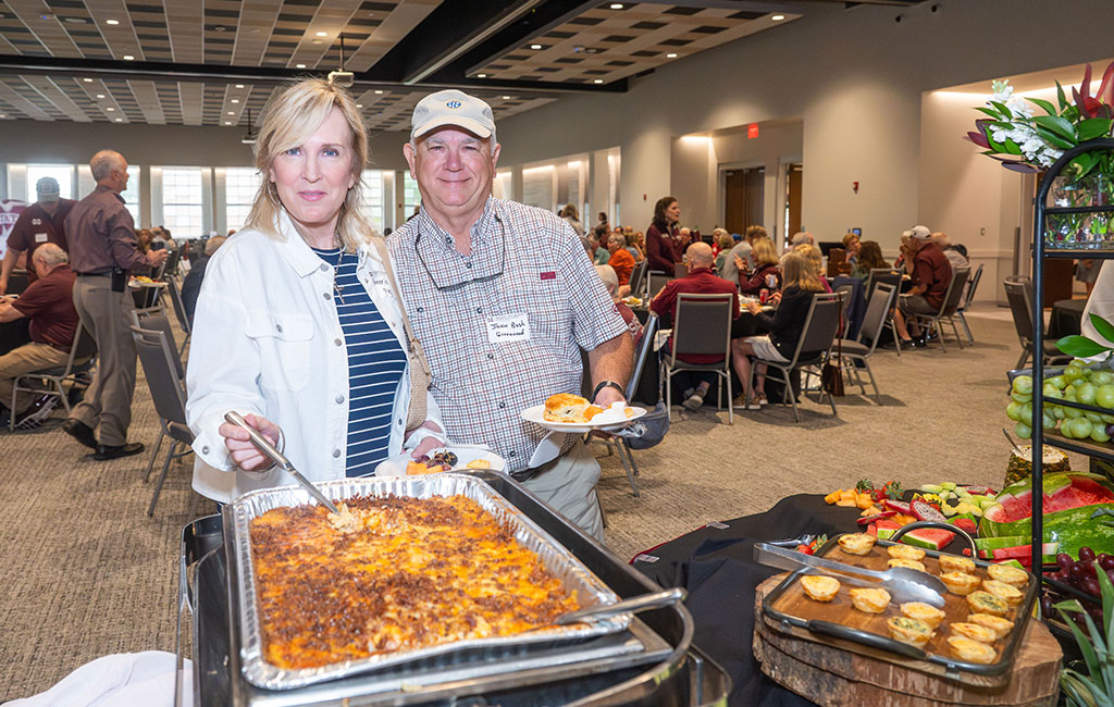 A woman serves casserole onto a man’s plate at a buffet table in a large, well-lit event room filled with people seated at round tables.