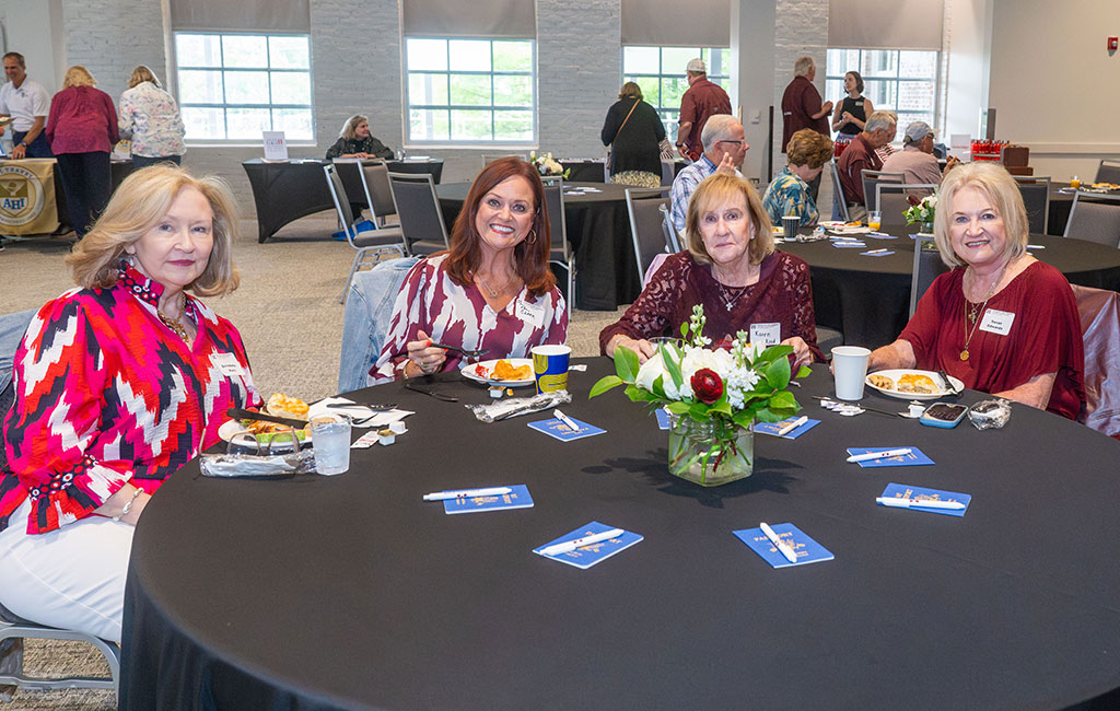 Four women sit at a round table with food and drinks in a large event room. A flower arrangement decorates the table. Several other people are visible in the background.