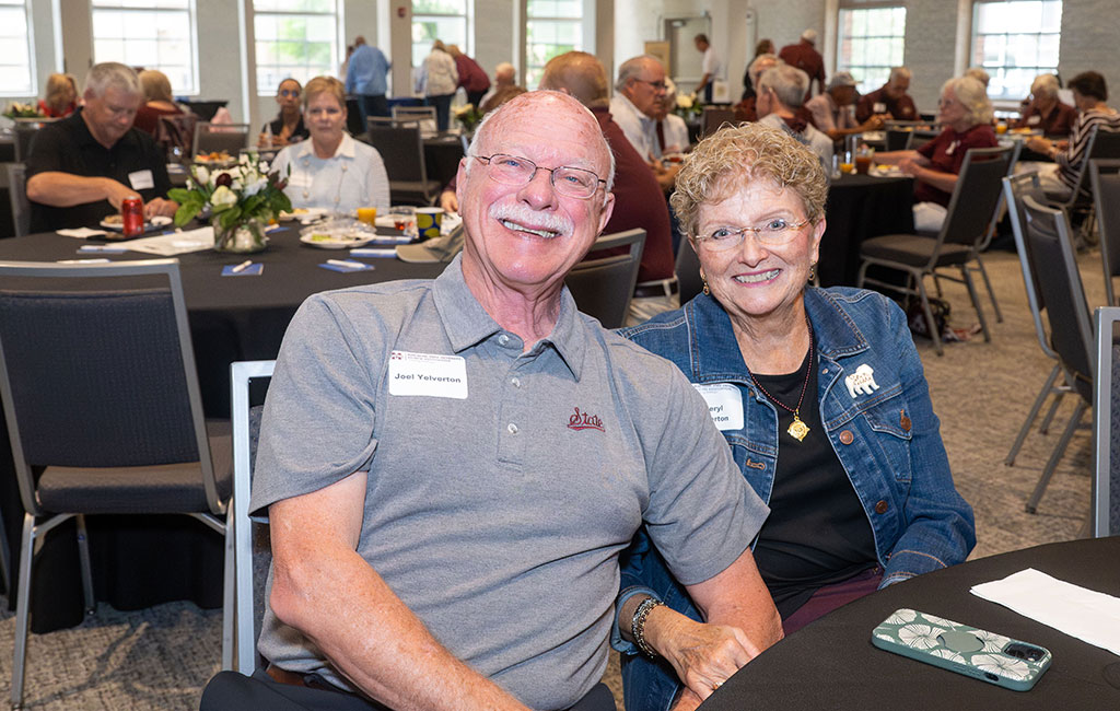 Two older adults smiling and sitting together at a round table during a social event in a large, well-lit room with other people in the background.