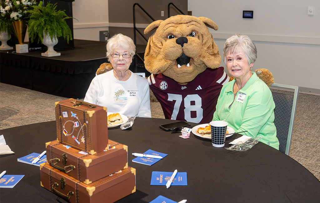 Two older women sit at a table with food, posing with a person in a bulldog mascot costume wearing a maroon jersey. A small suitcase decoration and brochures are on the table.