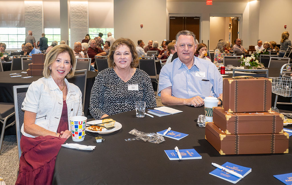 Three adults sit at a round table with food, drinks, and decorative boxes, attending an event in a banquet hall with other guests in the background.