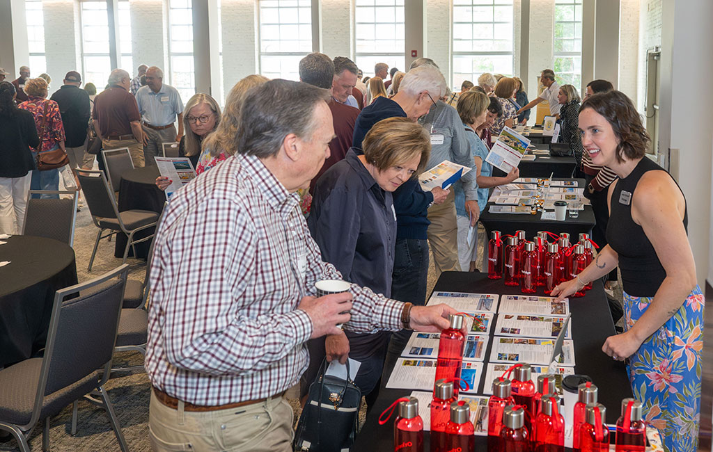 People gather around tables with pamphlets and red water bottles at an indoor event; a woman on the right speaks to attendees.