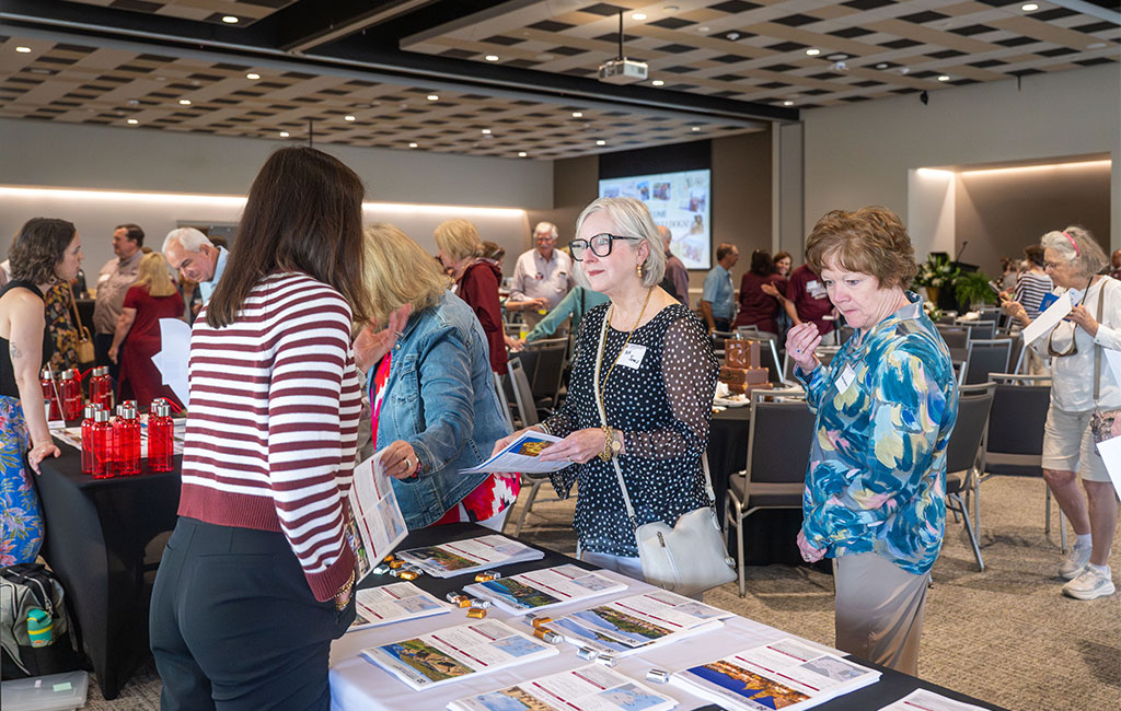 People stand around tables with pamphlets and information in a conference room, engaging in conversation at a daytime indoor event.