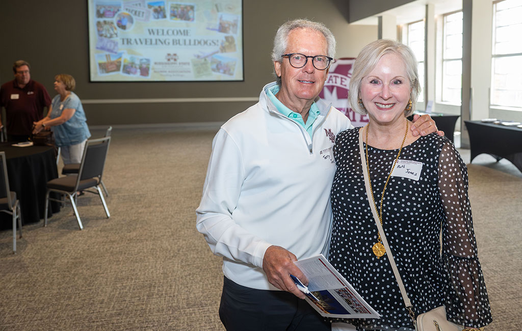 An older man and woman stand together, smiling at a social event. The man holds papers. Other attendees and a “Welcome Traveling Bulldogs” sign are visible in the background.