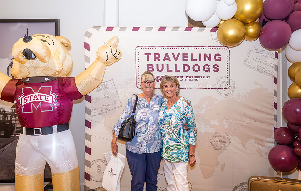 Two women stand smiling in front of a “Traveling Bulldogs” Mississippi State University backdrop, with an inflatable bulldog mascot and decorative balloons beside them.