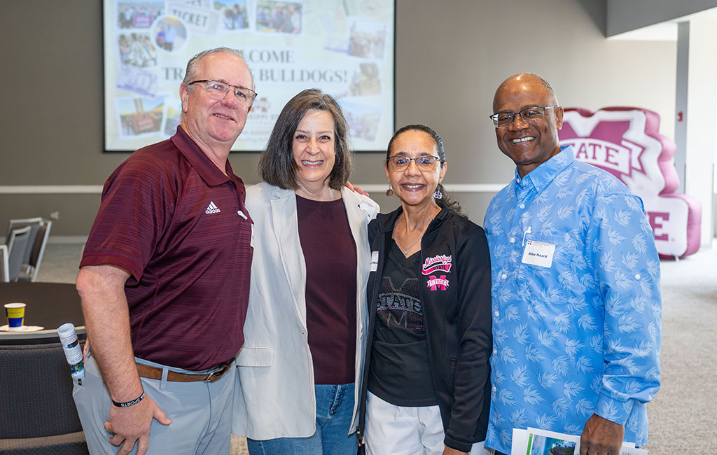 Four adults stand together indoors, smiling for a group photo; a presentation screen and university-themed decor are visible in the background.