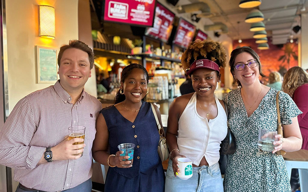 Four people stand together indoors, smiling and holding drinks in a lively bar or restaurant setting.