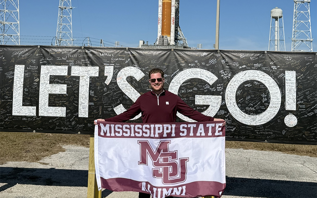 Person holding a Mississippi State University Alumni flag stands in front of a large “LET’S GO!” sign near a rocket launch site.