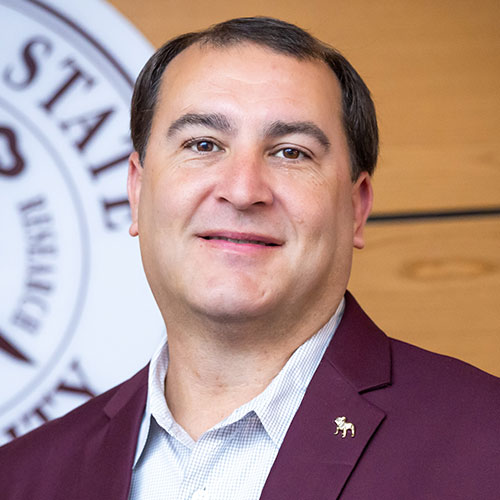 A man in a maroon blazer and light shirt stands in front of a partial seal or logo on a wooden background, facing the camera and smiling slightly.