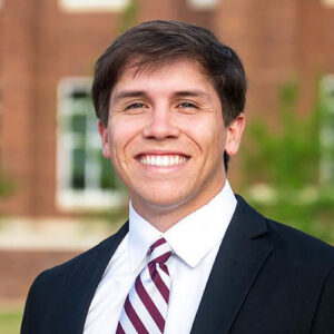 A man in a suit and striped tie smiles at the camera, standing outdoors in front of a blurred brick building and green trees.