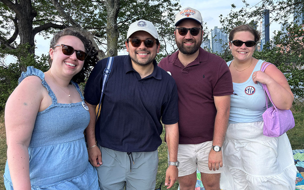 Four adults wearing sunglasses and casual summer clothing stand outdoors on grass, smiling at the camera, with trees and city buildings visible in the background.