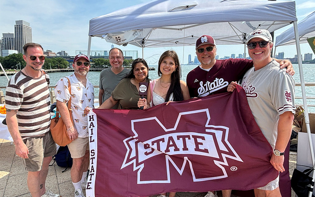Seven people stand under a tent by the water, holding a Mississippi State University “Hail State” flag and smiling at the camera.