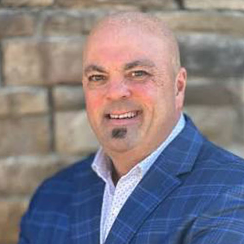 A bald man with a goatee wearing a blue plaid suit jacket and white shirt stands in front of a stone wall, smiling at the camera.