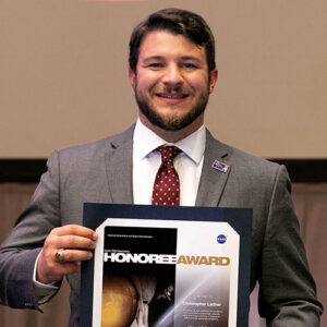 Man in a suit holds a NASA Honoree Award certificate and smiles at the camera.