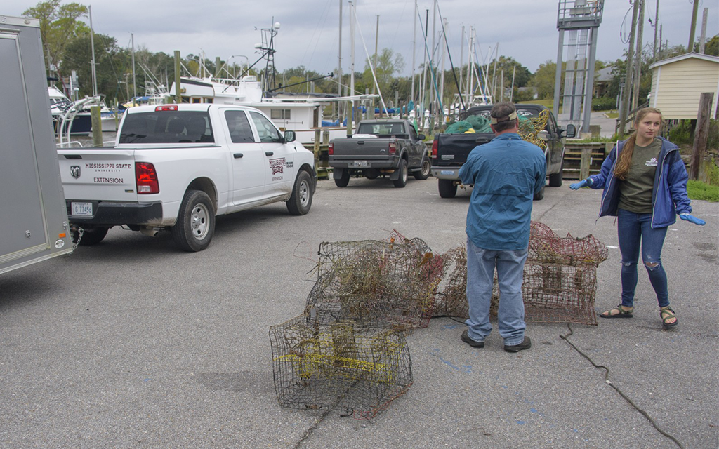 Two people stand near several wire crab traps on a dock parking lot, with trucks, boats, and docks visible in the background.
