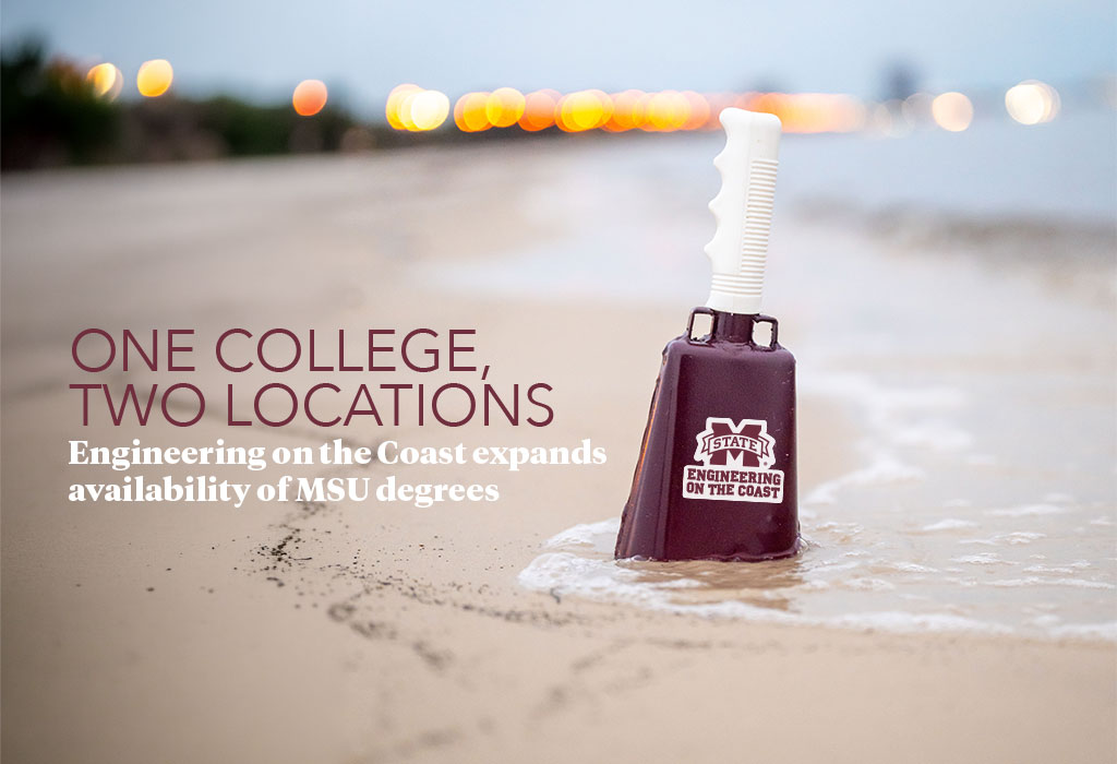 A maroon cowbell with “Engineering on the Coast” printed on it stands upright in wet sand near the shoreline, with blurred lights in the background.