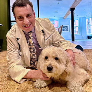 A veterinarian in a white coat kneels on the floor, smiling and holding a fluffy light-colored dog in a bright room.
