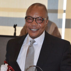 A man in a suit and tie smiles while holding an award and a bouquet of flowers indoors, with blurred people and a microphone stand in the background.