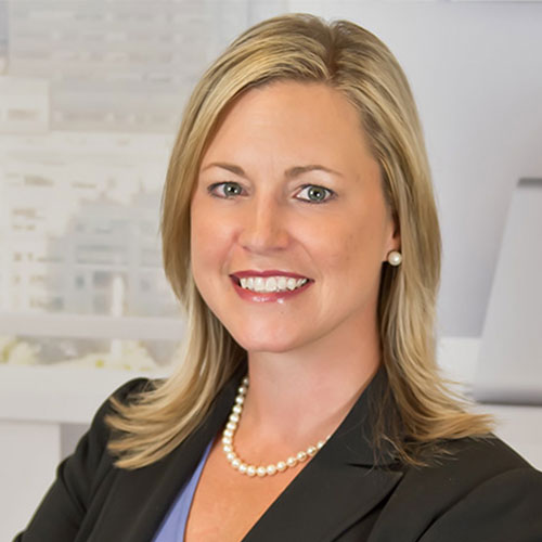 A woman with blonde hair wearing a black blazer, pearl necklace, and earrings smiles at the camera against a blurred office background.
