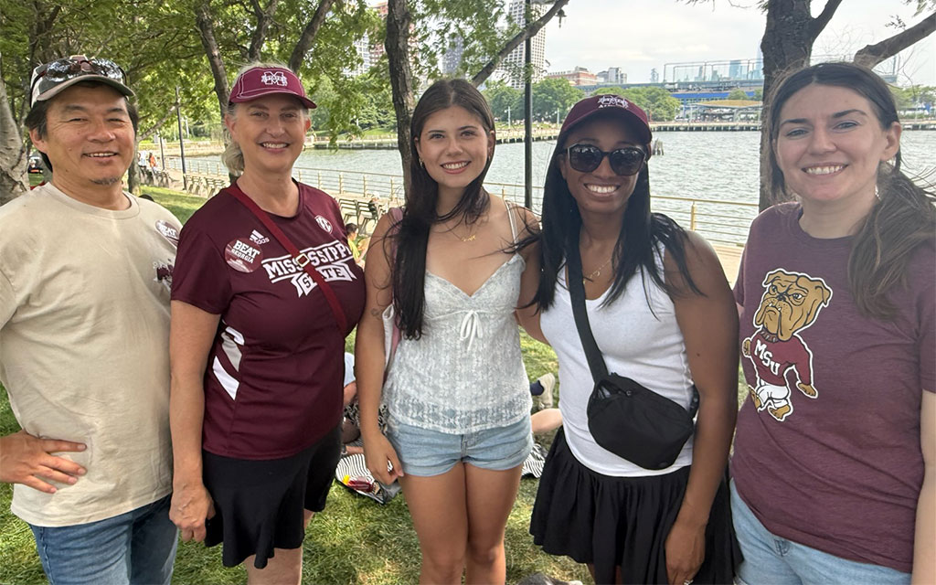 Five people stand smiling outdoors by a riverside, with trees and city buildings in the background. Some wear Mississippi State University shirts and hats.
