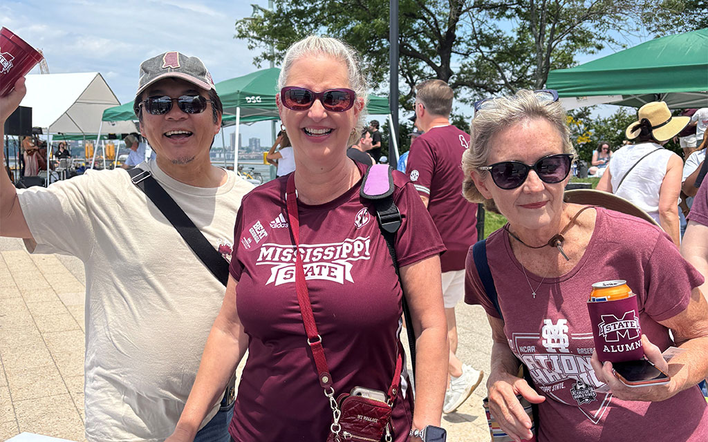 Three adults wearing Mississippi State shirts and sunglasses stand outdoors at an event, smiling for the camera with tents and other people visible in the background.