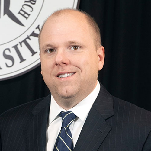A man in a pinstripe suit and striped tie stands in front of a university seal, smiling at the camera.