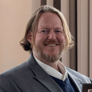 Man with shoulder-length blond hair and beard wearing a gray suit jacket, white shirt, and sweater, standing indoors in front of beige and brown vertical panels.