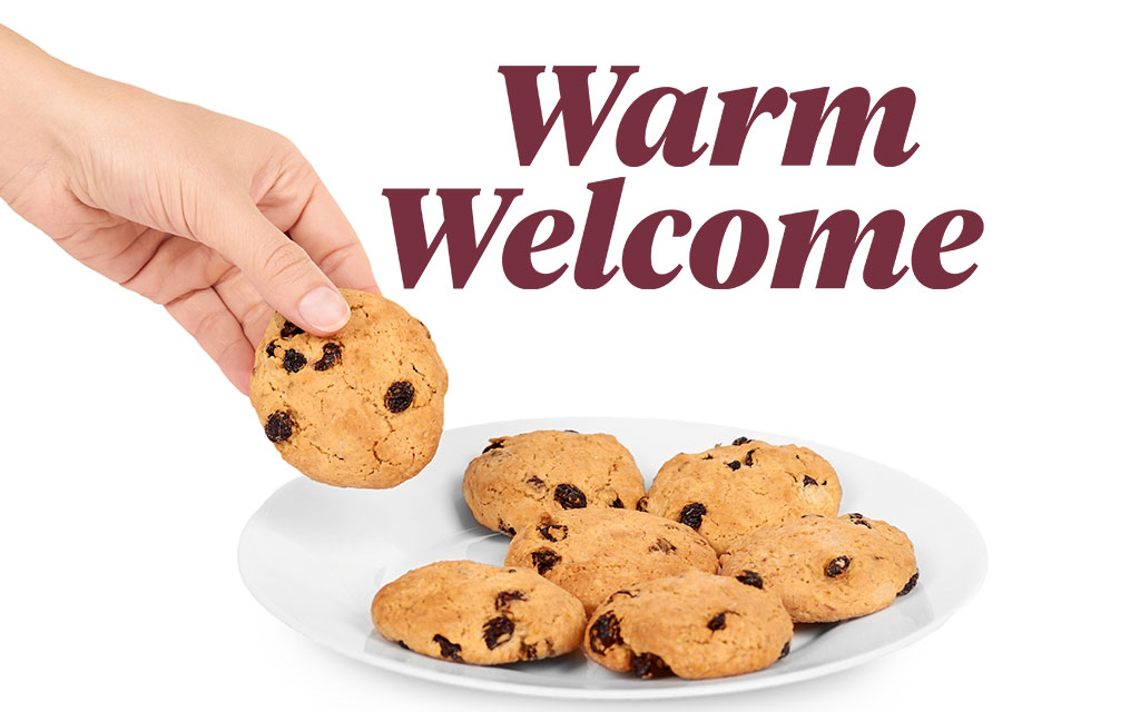 A hand picks up a raisin cookie from a plate of cookies, with the words “Warm Welcome” in large text above.